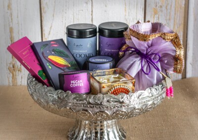 Floral Art presents A decorative platter holds two tins of cookies, chocolate bars, a packet of pecan coffee, and several small gift-wrapped items, perfect for any occasion or to complement exquisite flower jewelry for bride.