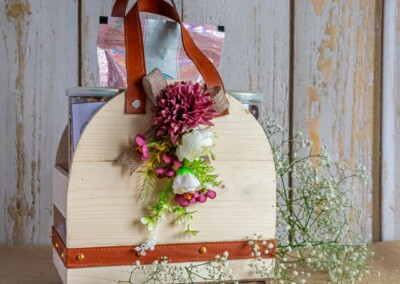Floral Art presents A wooden gift basket with leather straps, adorned with floral bridal jewelry, containing various items inside. It is placed on a wooden surface and surrounded by sprigs of white baby's breath flowers.