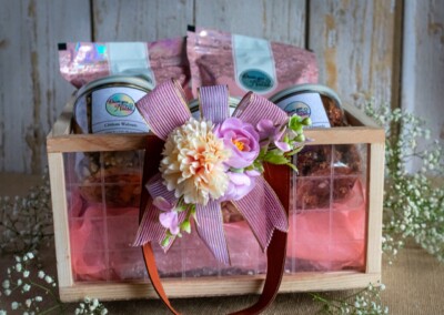 Floral Art presents A wooden gift basket with a pink ribbon and floral decoration containing packaged food items, beautifully placed on a textured surface with small flowers scattered around, making it an ideal setting reminiscent of delicate floral bridal jewellery.
