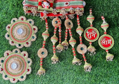 Floral Art presents A decorative set laid on grass includes two round pieces, one long horizontal piece with intricate designs, and several hanging ornaments in red inscribed with Hindi text. Perfect for a wedding varmala or floral jewelry for the bride.