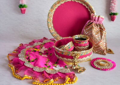 Floral Art presents A pink decorative set featuring a round tray, fabric pouch, embellished dress, and matching accessories displayed on a light background with floral garlands above. The addition of exquisite flower jewelry for the bride enhances the elegance of this charming ensemble.