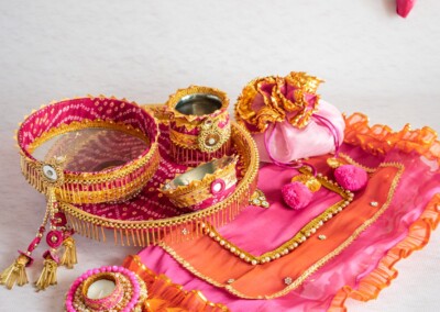 Floral Art presents A Karva Chauth thali set with decorated sieve, pots, ornaments, and a colorful cloth is beautifully displayed on a white surface, complemented by flower jewelry for haldi and pink woven garlands above.