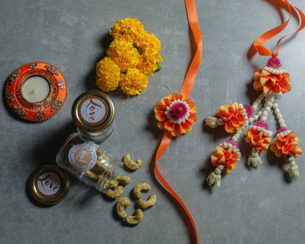 Floral Art presents A decorative candle, marigolds, jars of cashews labeled 'Love', and a floral garland with orange ribbon are arranged on a gray surface.