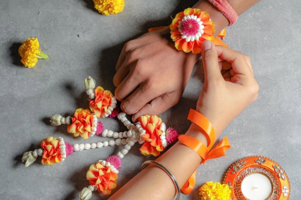 Floral Art presents Two hands with floral bracelets made of marigold petals and beads, with more floral decor and a lit candle on a gray surface. The text "floral art" is overlaid on the image.