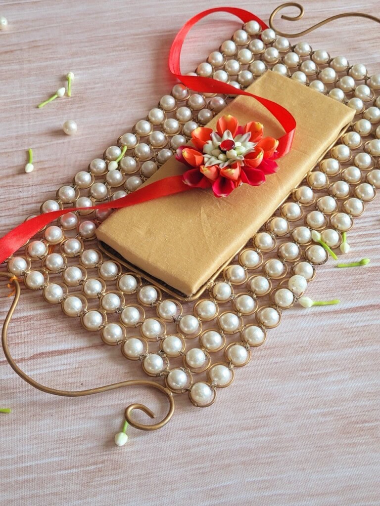 Floral Art presents A gold envelope with a red and white flower on top placed on a pearl studded rectangular mat with a red ribbon, against a wooden background.