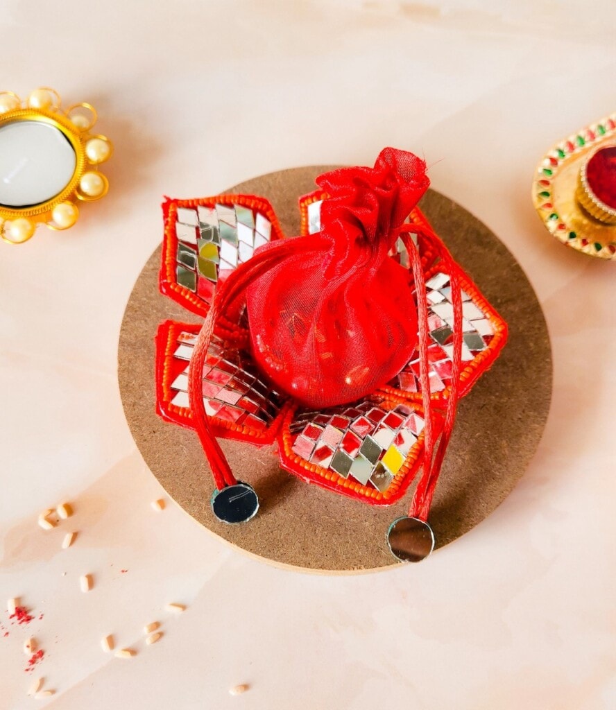 Floral Art presents A small red decorative pouch with shiny mirrored accents on a brown base, surrounded by traditional Indian decorative items, including a small lamp and candle.