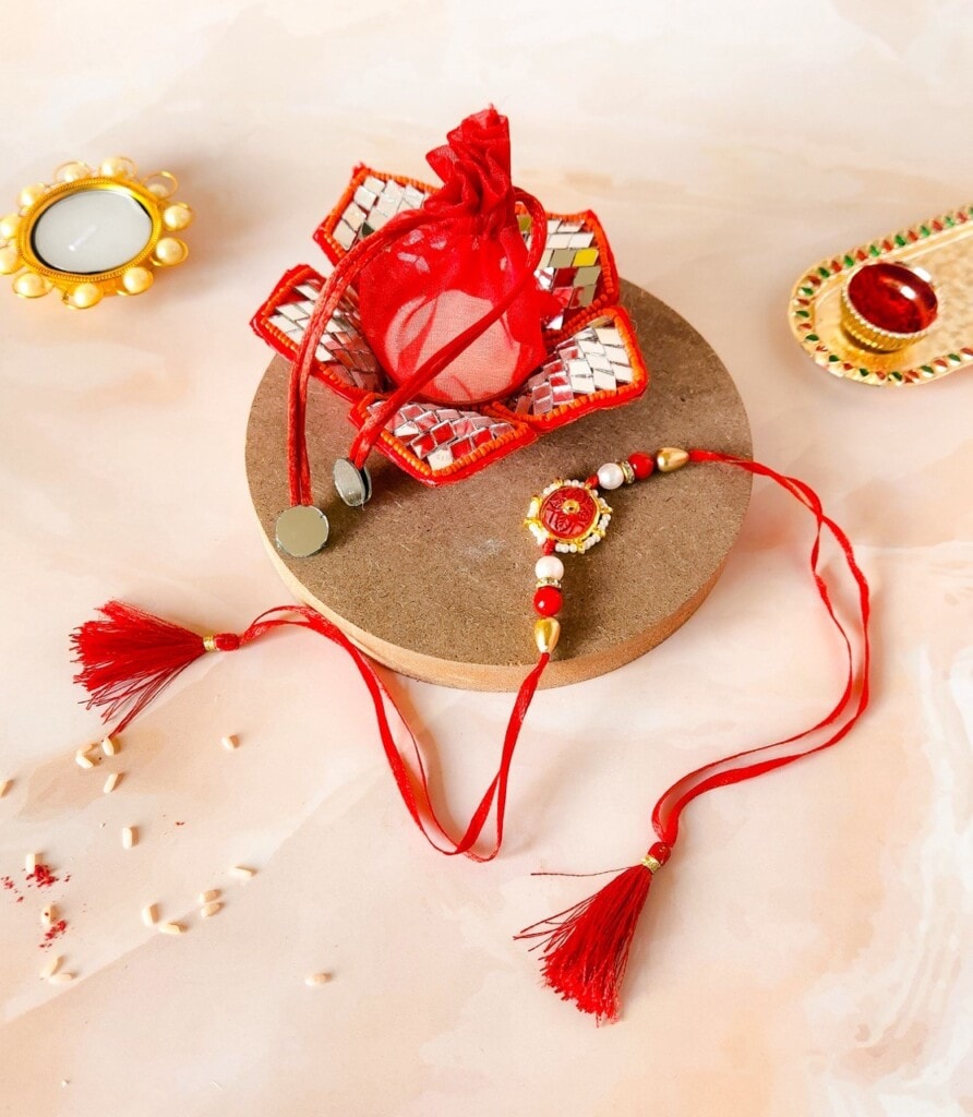 Floral Art presents A decorative Rakhi with red and gold details is displayed with a small pouch, a lit candle, and traditional festive items on a flat surface.