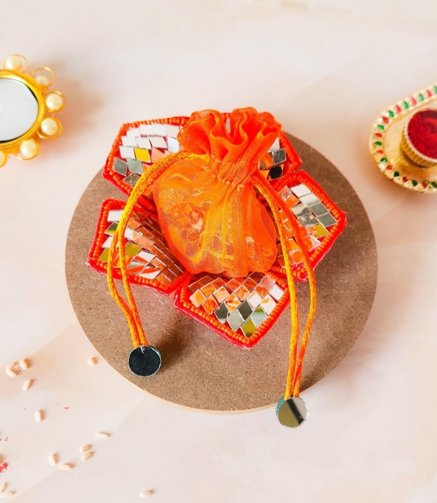 Floral Art presents An orange drawstring pouch is placed on a beaded circular coaster on a light surface, with decorative items including a small mirror and a dish seen in the background.