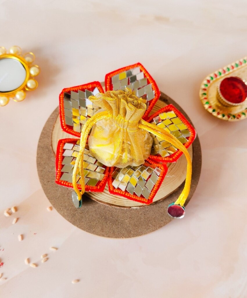 Floral Art presents A decorative arrangement featuring a small yellow drawstring pouch, surrounded by five reflective, red bordered items, placed on a wooden base with a candle and a decorative holder nearby.