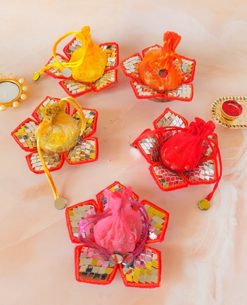 Floral Art presents Five colorful potlis are displayed, each placed on a decorative flower shaped base with mosaic designs. The potlis vary in colors: yellow, orange, red, and pink. A decorative mirror and bindi are visible.