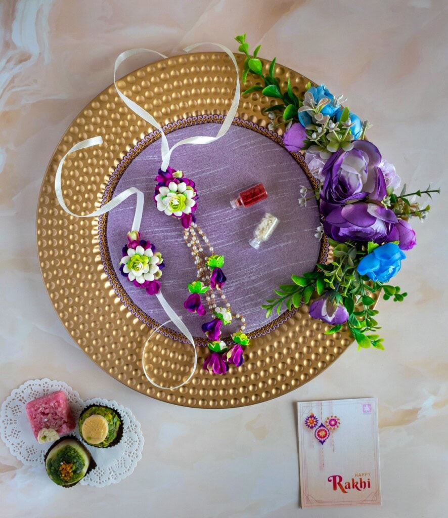 Floral Art presents A decorated plate with purple and green floral accessories, a small bottle, and a notecard, next to a plate with sweets and a card reading "Rakhi.
