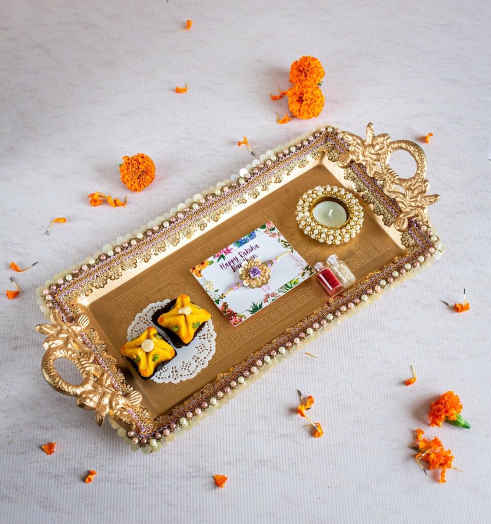 Floral Art presents A decorated tray with marigold flowers, sweets, a candle, a small vial, and a card. The tray has ornate handles and a decorative border.