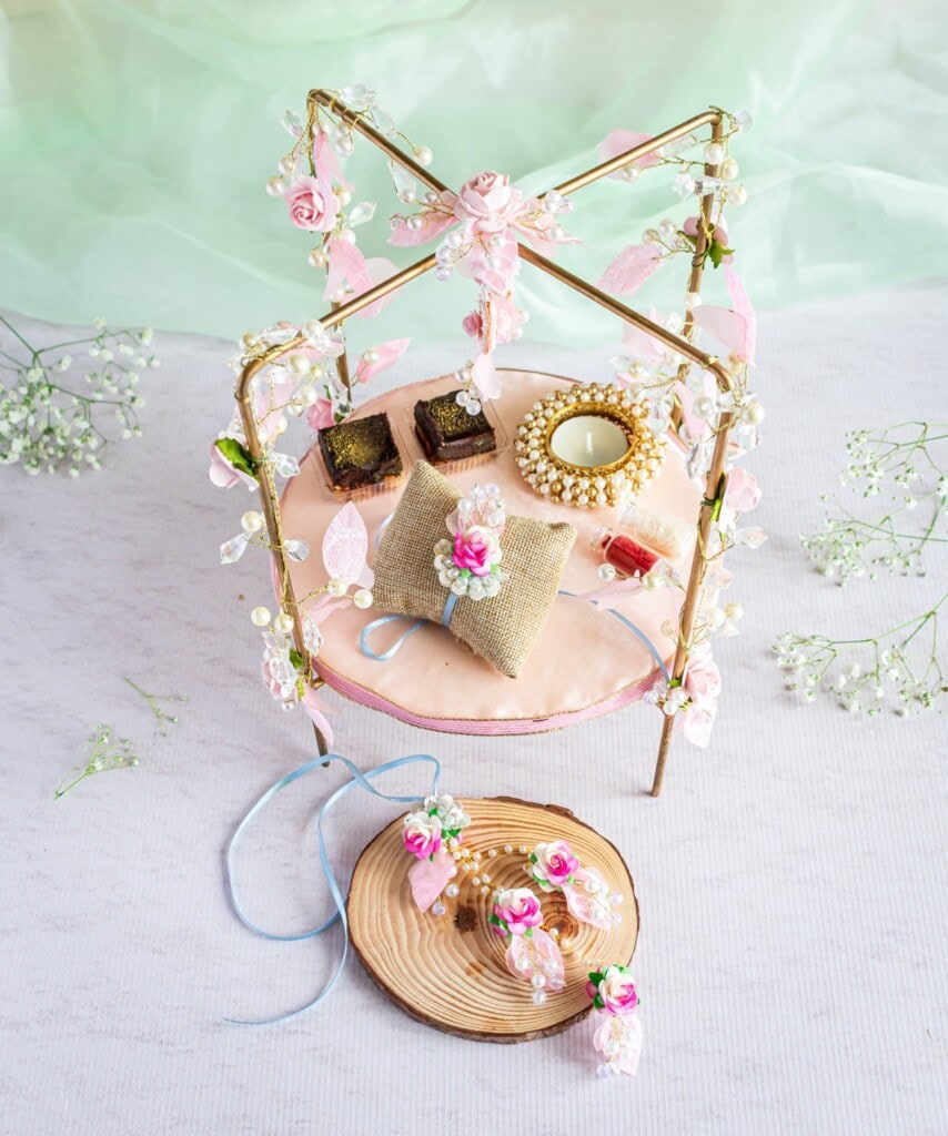 Floral Art presents A mini decorative setup with a small table adorned with flowers, ribbons, and a lace canopy. On the table are two cakes, a golden frame with a mirror, a burlap pillow, and a wooden slice with bouquets.