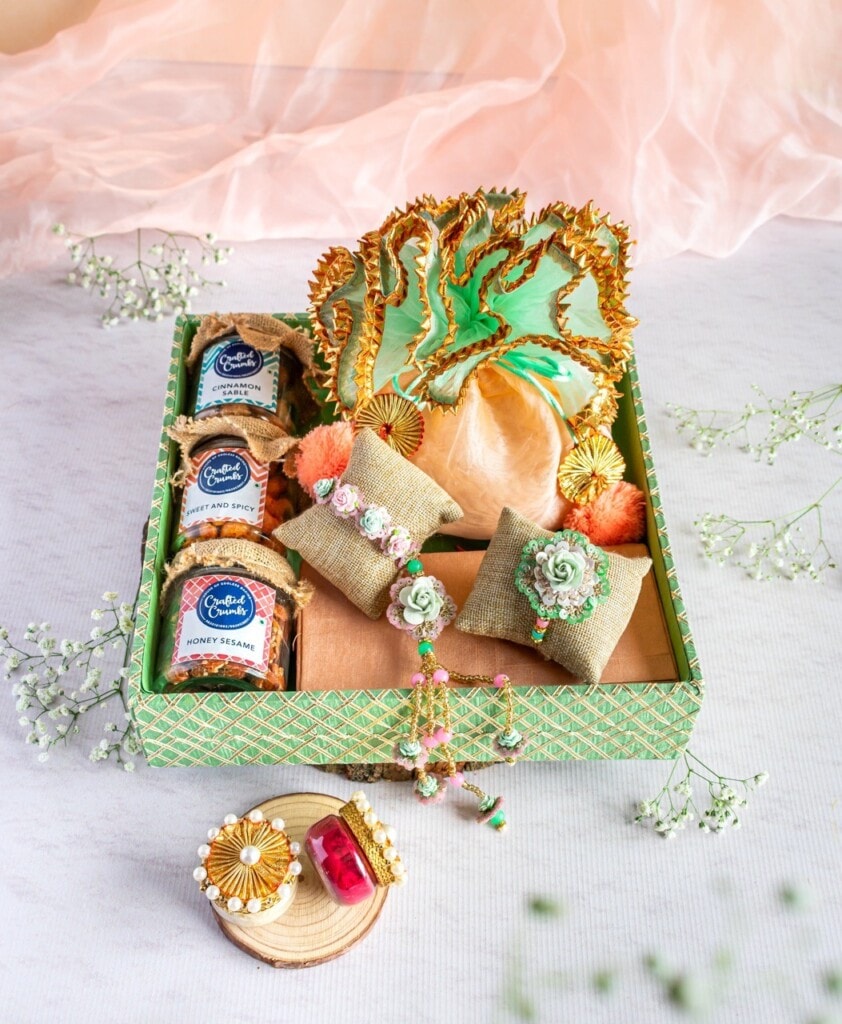 Floral Art presents A decorative gift box containing traditional sweets, small fabric cushions with jewelry, and ornate green leaves on top, placed on a white surface with a pink sheer fabric in the background.