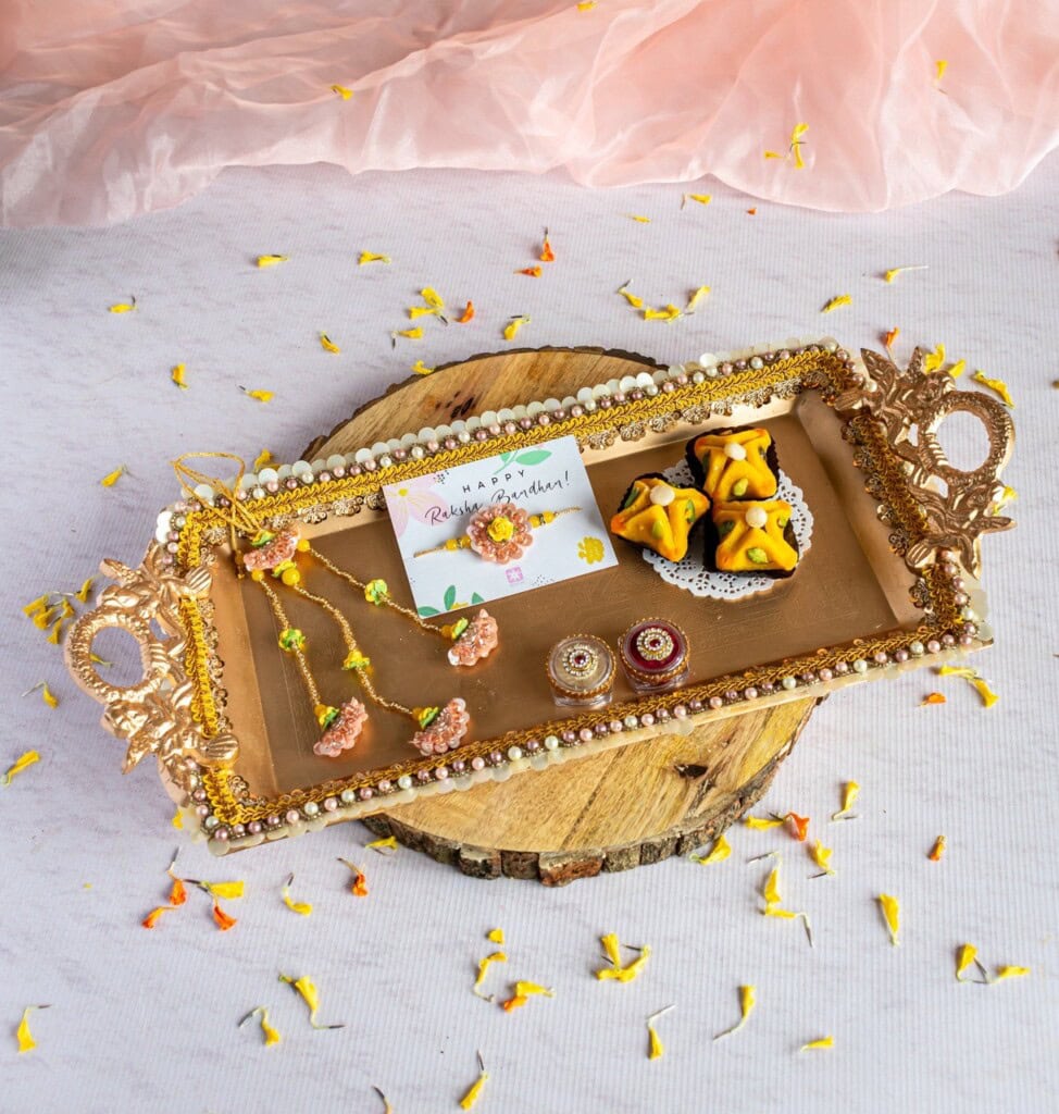 Floral Art presents A decorative tray with Rakhi, a greeting card, sweets, and a small figurine, surrounded by scattered flower petals on a wooden surface.