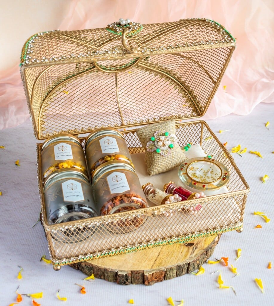 Floral Art presents An open, ornate wire gift box contains various colorful items, including jars, a small pouch, rolled items, and a decorative piece, all placed on a wooden base against a soft pink backdrop.