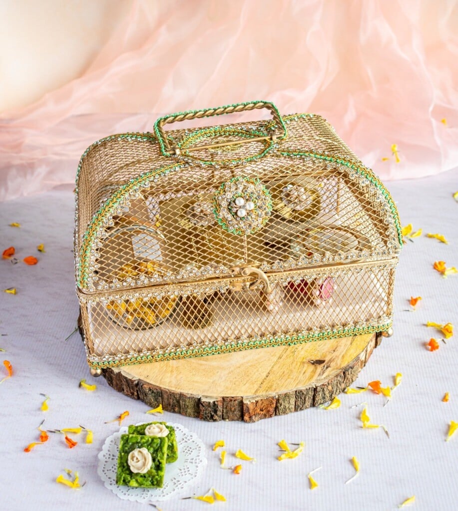 Floral Art presents A decorative golden cage with intricate details and a handle sits on a wooden slice. Inside the cage are food items. Flower petals are scattered around, and a green dessert is placed nearby.