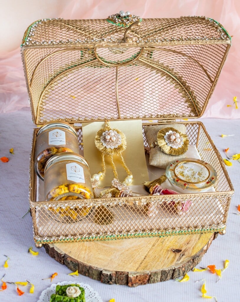 Floral Art presents A golden wire mesh gift basket contains various items including jars of snacks, a rolled paper, jewelry, and decorative accessories, set on a wooden slab with flower petals around.