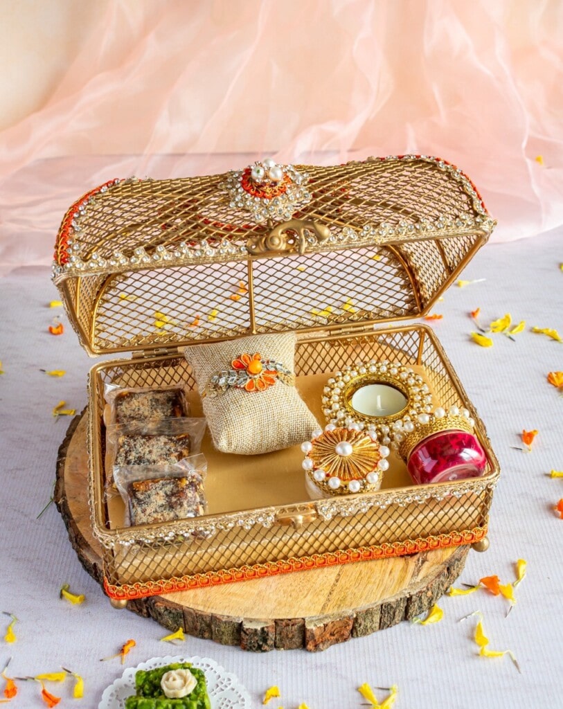 Floral Art presents A decorative open metal gift box with intricate designs contains wrapped sweets, a small candle, a pouch, and a red container. The box is placed on a wooden slice with scattered flower petals around.