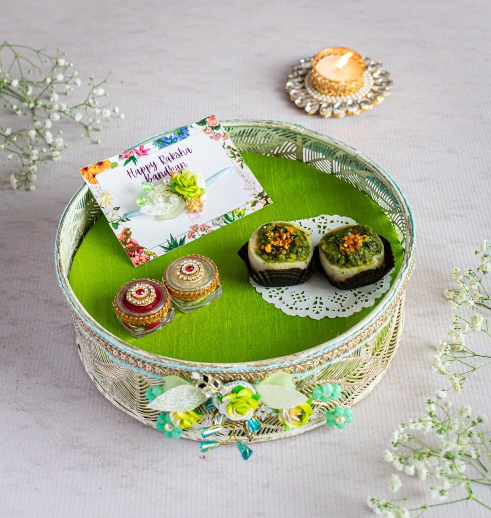 Floral Art presents A decorative tray with two sweets, small containers of kumkum and rice, a lit candle, and a "Happy Raksha Bandhan" card on a green base, surrounded by white flowers.
