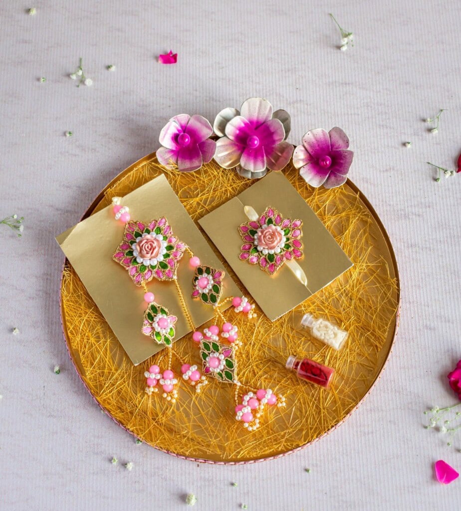 Floral Art presents A gold colored platter with intricate floral jewelry pieces, decorative flowers, and small vials of red and white substances on a textured surface.