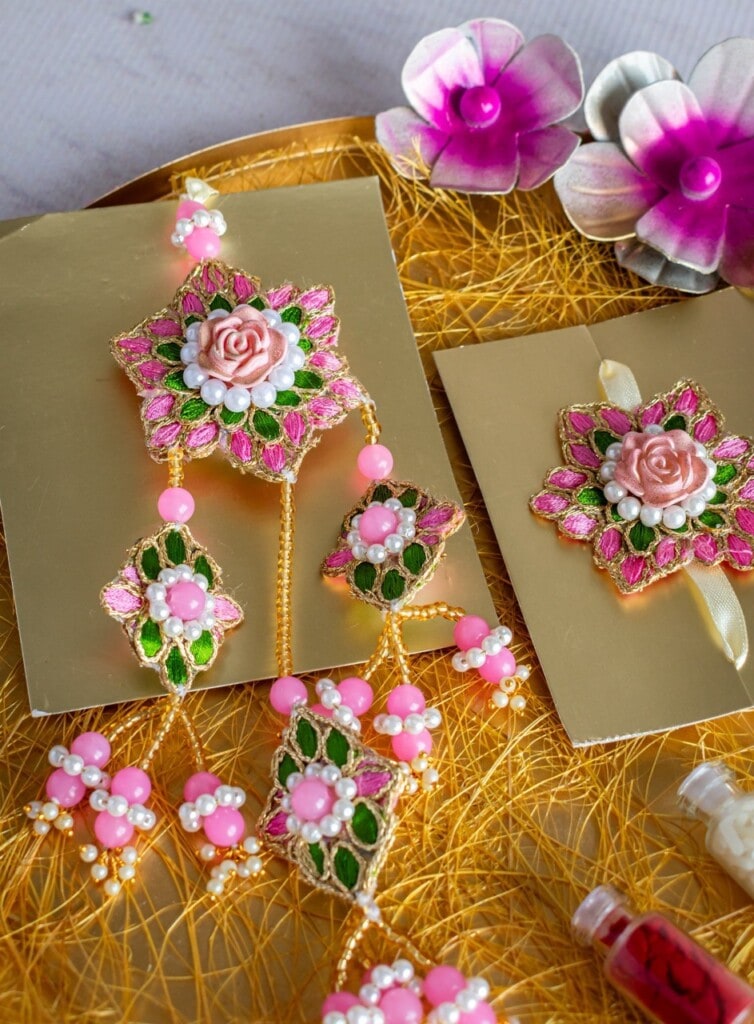 Floral Art presents A decorative jewelry set with floral designs, pink and green beads, and a central rose motif is displayed on a golden textured surface next to artificial flowers.