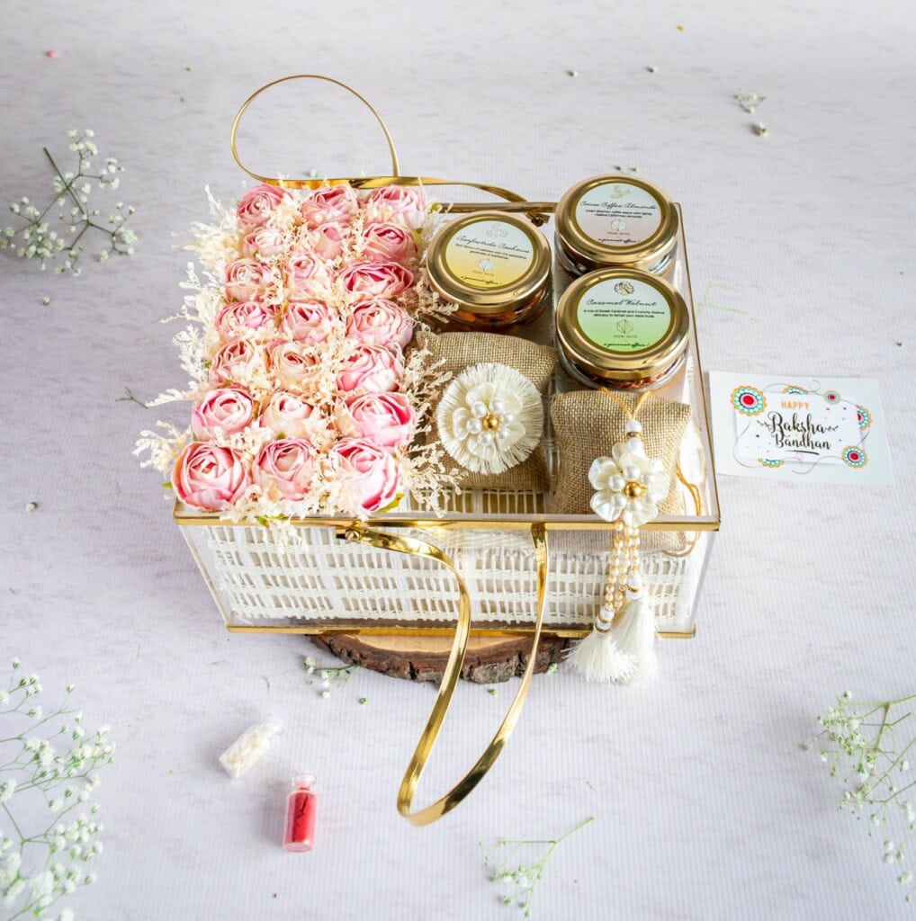 Floral Art presents A decorative gift basket with jars of preserves, a bunch of light pink roses, and a couple of wrapped items. The basket is adorned with gold accents and accompanied by a Raksha Bandhan card.