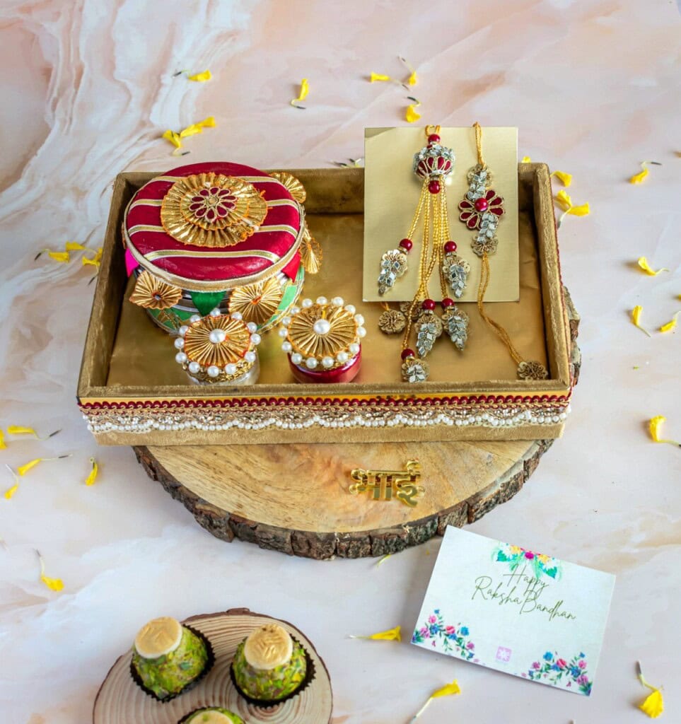 Floral Art presents A decorative tray contains traditional jewelry, a round ornate box, and a card. The tray is placed on a wooden base beside a bowl of sweets, with scattered petals on a marble surface.