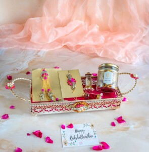 Floral Art presents A decorative tray with a Rakhi set, including pink and gold rakhis, a jar of sweets, and an envelope. A greeting card with "Happy Raksha Bandhan" is displayed in front. Pink fabric and petals are in the background.