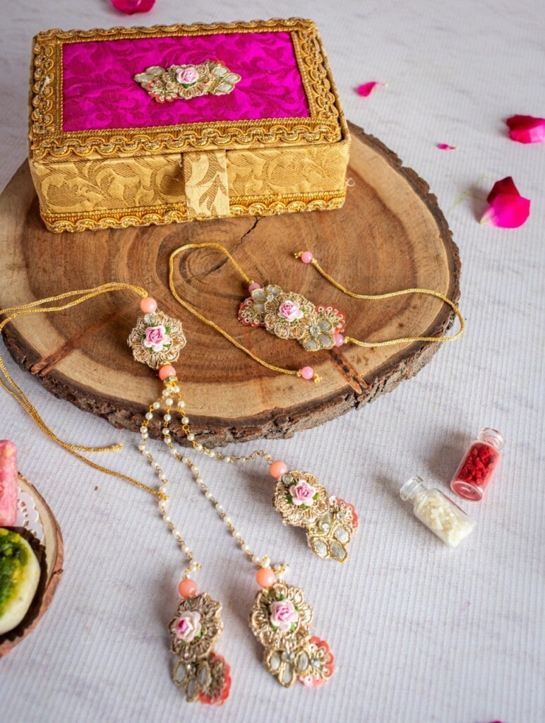 Floral Art presents A gold and pink jewelry set is displayed on a wooden platter alongside a decorative box, rose petals, and small vials of colored powder.