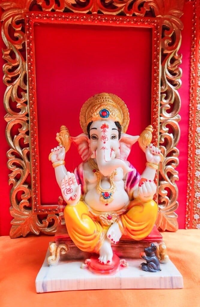 Floral Art presents A statue of Ganesha in vibrant colors is placed in an ornate red and gold frame. Ganesha, the Hindu deity, is shown with four arms holding symbolic items and a small mouse at his feet.