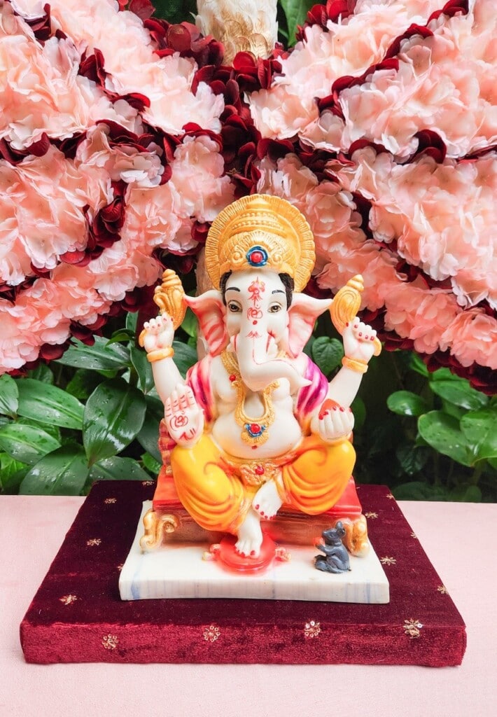 Floral Art presents A statue of the Hindu deity Ganesha, adorned with vibrant colors, is seated on a decorated platform in front of pink and red flowers.