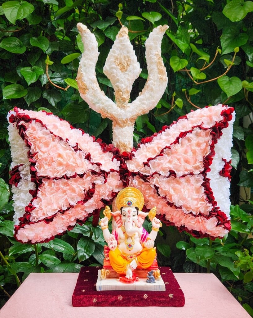 Floral Art presents An idol of Lord Ganesha is placed in front of a large trident shaped floral arrangement with pink and white flowers, set against a backdrop of green foliage.