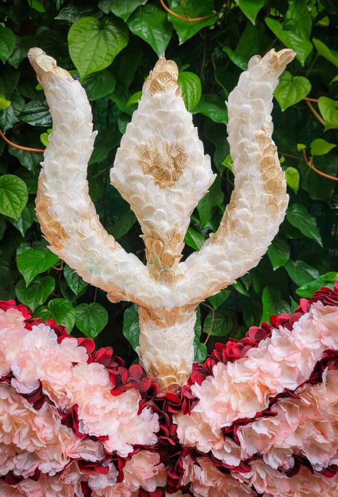 Floral Art presents A white trident shaped floral sculpture is set against a background of green leaves, with pink and red flowers arranged at its base.