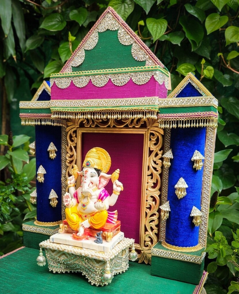 Floral Art presents A decorated idol of a seated Ganesh, positioned in front of an ornate, colorful backdrop with a green and leafy background.