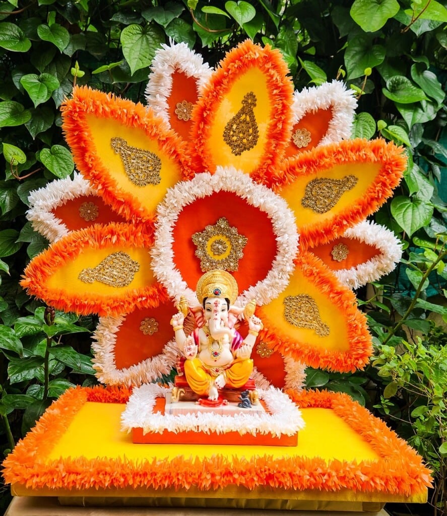 Floral Art presents A statue of Ganesha is placed on a decorated altar with a vibrant orange and yellow floral backdrop, surrounded by greenery.