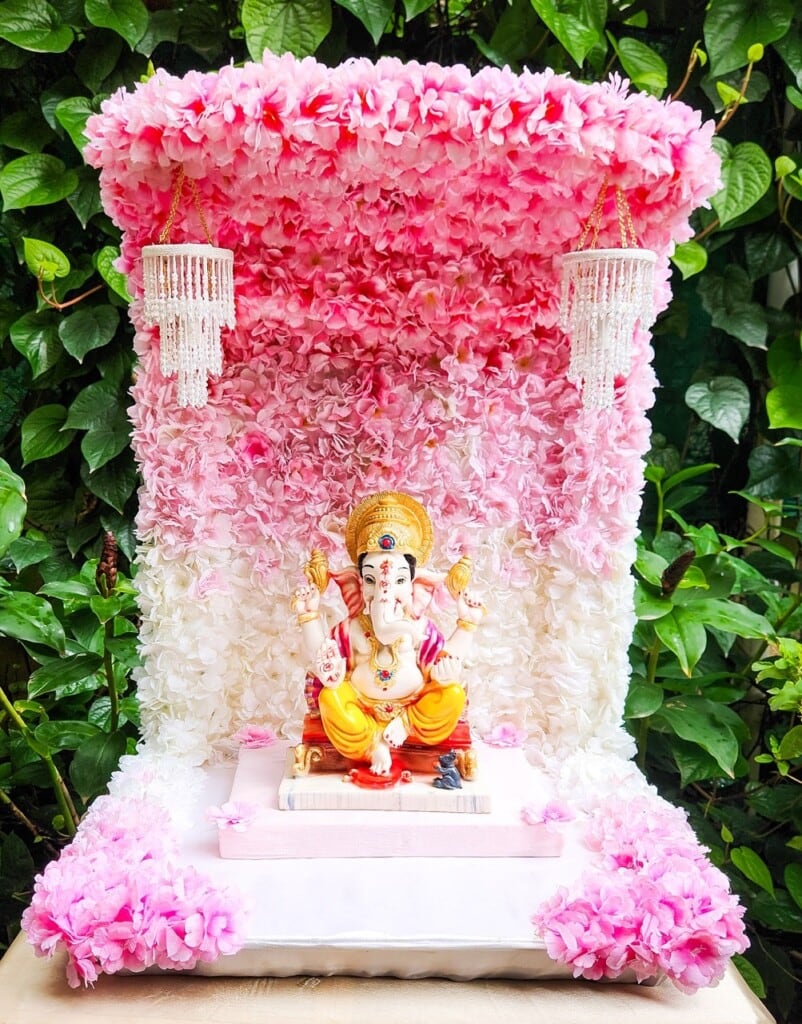 Floral Art presents A statue of Lord Ganesha is placed on a platform adorned with pink and white flowers, with two hanging ornaments on either side, set against a backdrop of green foliage.