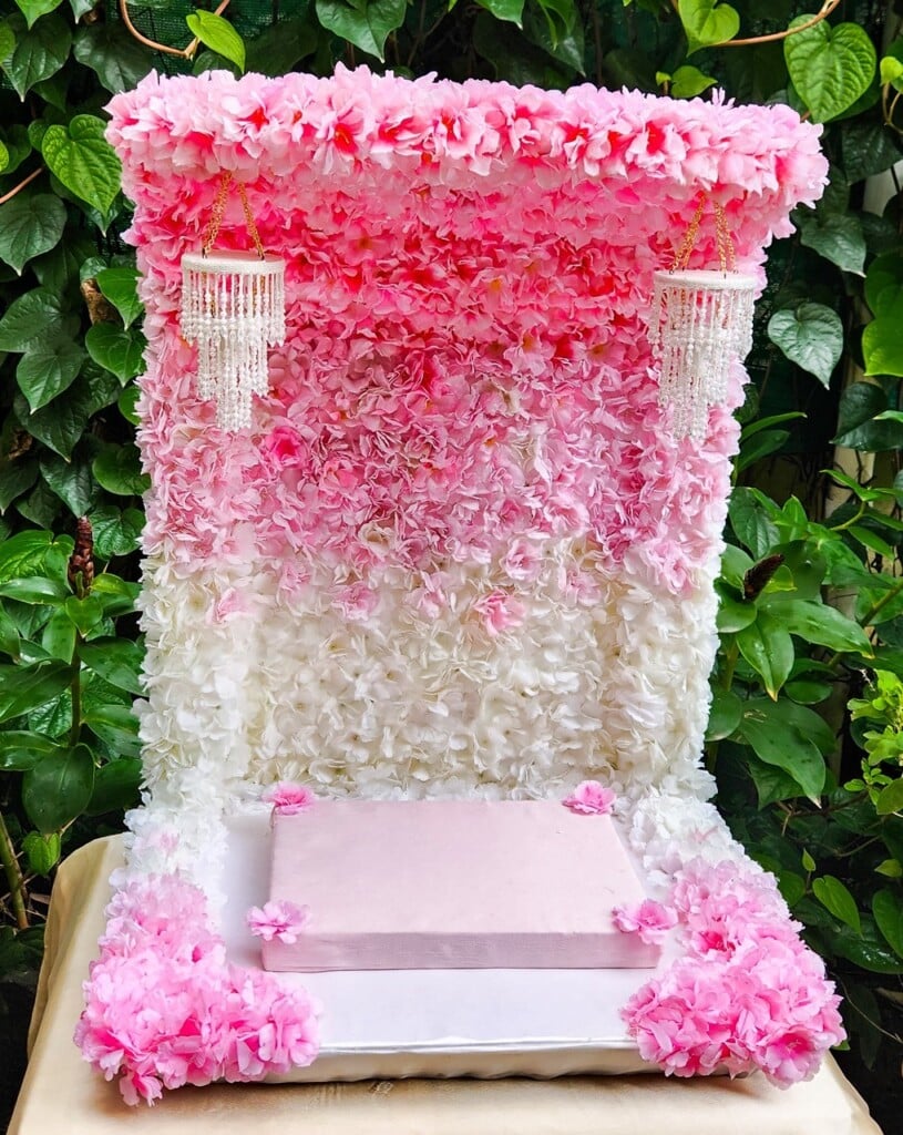 Floral Art presents A rectangular flower wall adorned with pink and white flowers, featuring two hanging chandeliers, and a table with a pink cloth at the bottom.