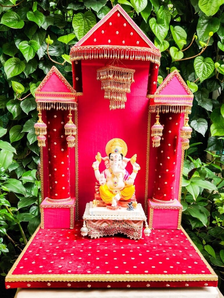 Floral Art presents A statue of Ganesha is placed on a decorative red platform with gold accents, surrounded by an ornate red and gold three tower backdrop; green foliage is visible in the background.