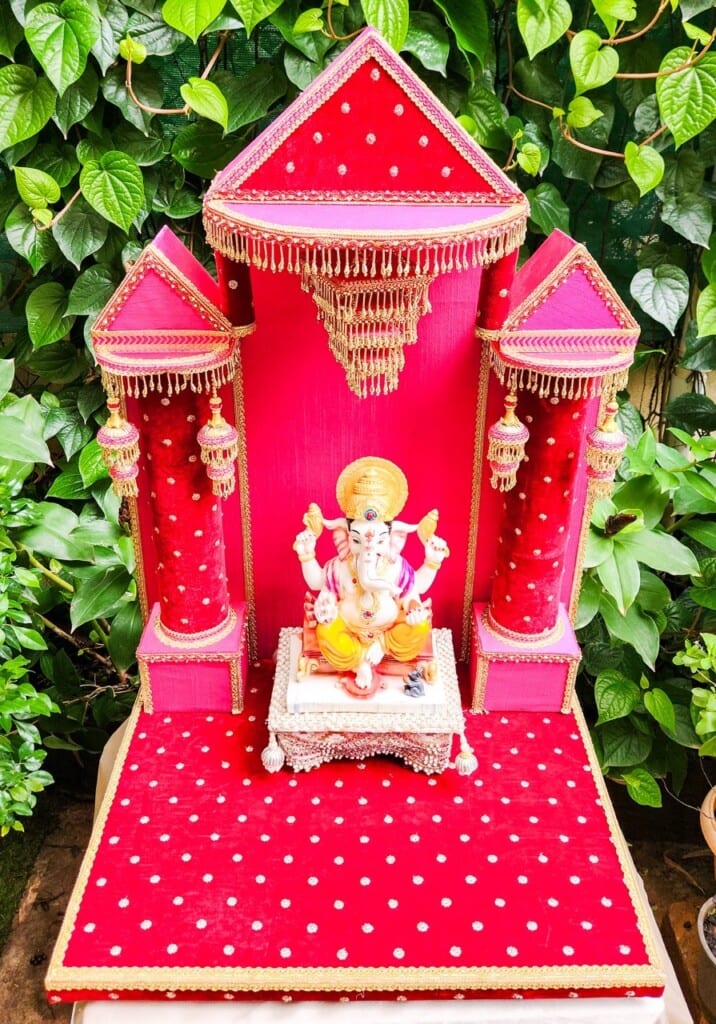 Floral Art presents A red decorated altar with golden accents, featuring an idol of Lord Ganesha seated on a silver pedestal, surrounded by green leaves.
