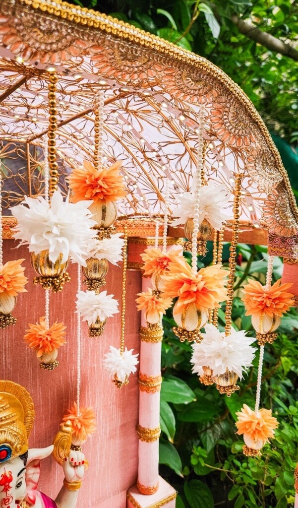 Floral Art presents Close up of an ornate decorative canopy featuring hanging orange and white floral garlands, with part of a deity statue partially visible in the bottom left corner. Lush greenery in the background.