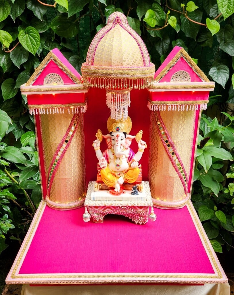 Floral Art presents A statue of Lord Ganesha is placed on a decorated platform with a pink backdrop and green leaves in the background.