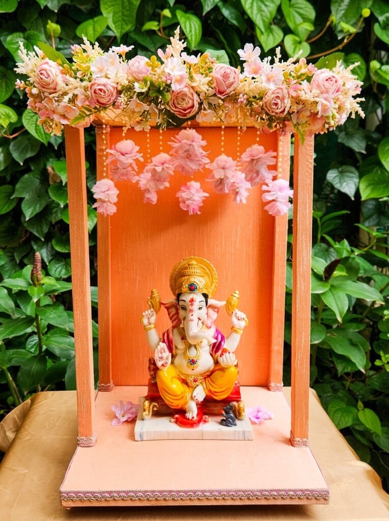 Floral Art presents An ornate Ganesha idol is seated under a decorated canopy with pink flowers and green foliage in the background.