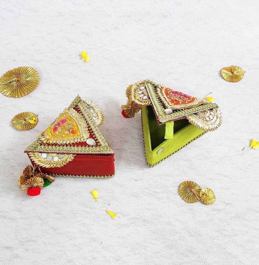 Floral Art presents Two intricately decorated, triangular dome gift boxes, one red and one green, on a light surface, surrounded by small golden decorations and yellow flower petals.