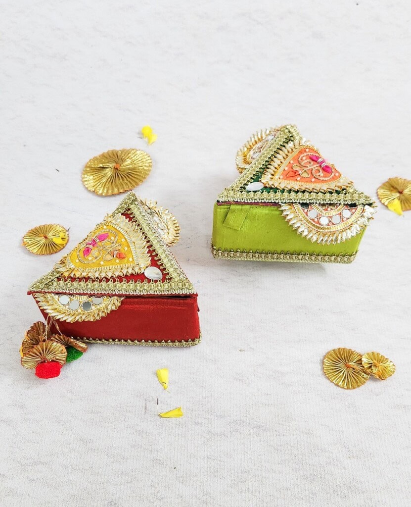 Floral Art presents Two decorative triangular boxes, one red and one green, adorned with intricate golden designs, sit on a white surface. Surrounding them are small golden decorative items and yellow flower petals.