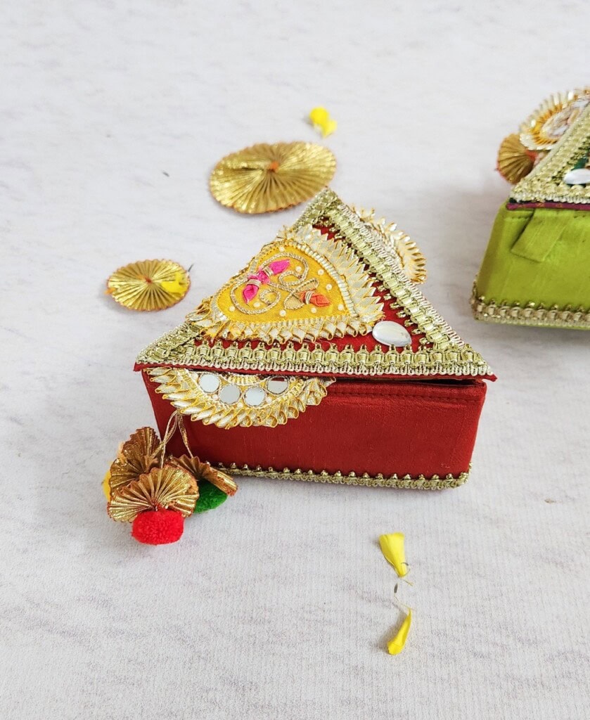 Floral Art presents Decorative red and green triangular gift boxes with intricate gold patterns and embellishments, placed on a white surface with some scattered decorative elements.
