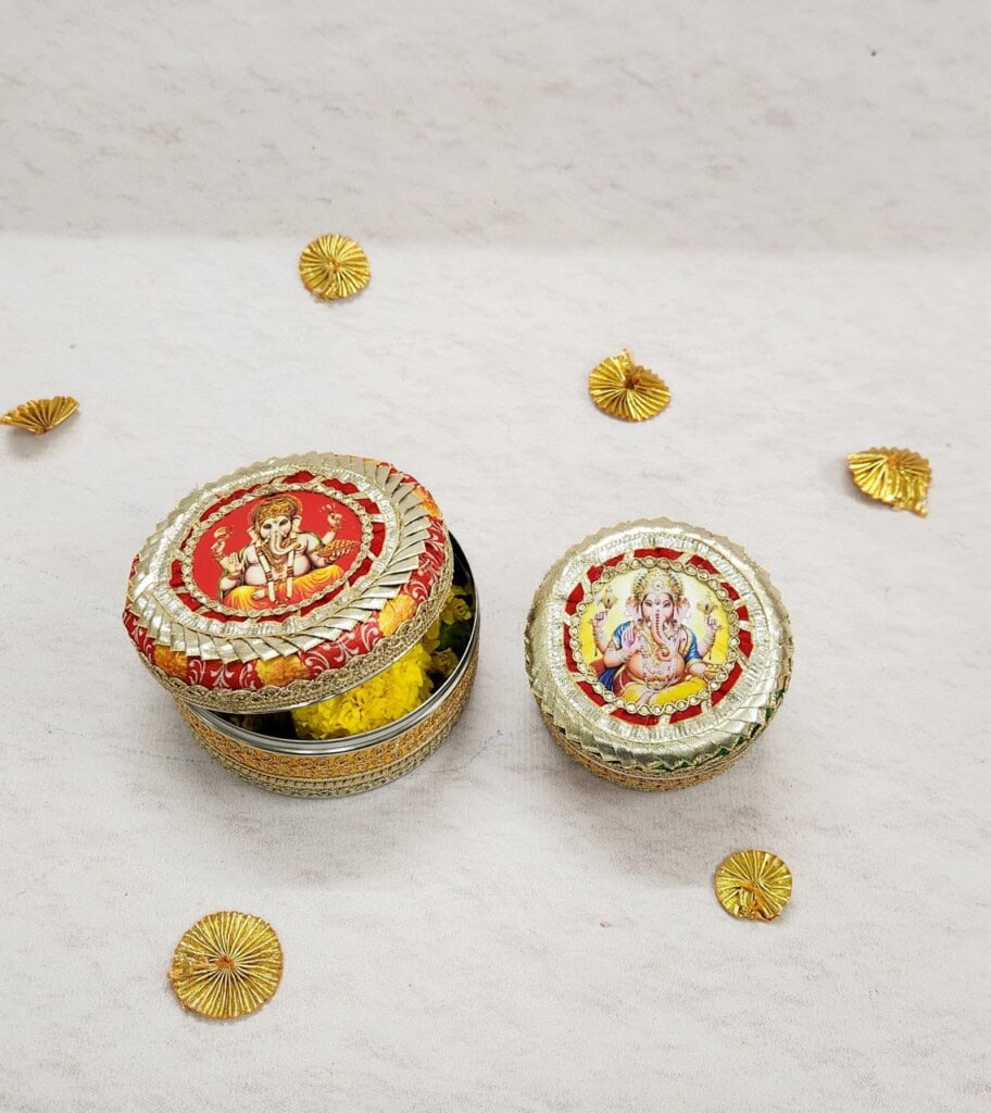 Floral Art presents Two ornate, metallic containers with illustrated lids featuring Hindu deities, placed on a white surface with small decorative gold elements scattered around them.