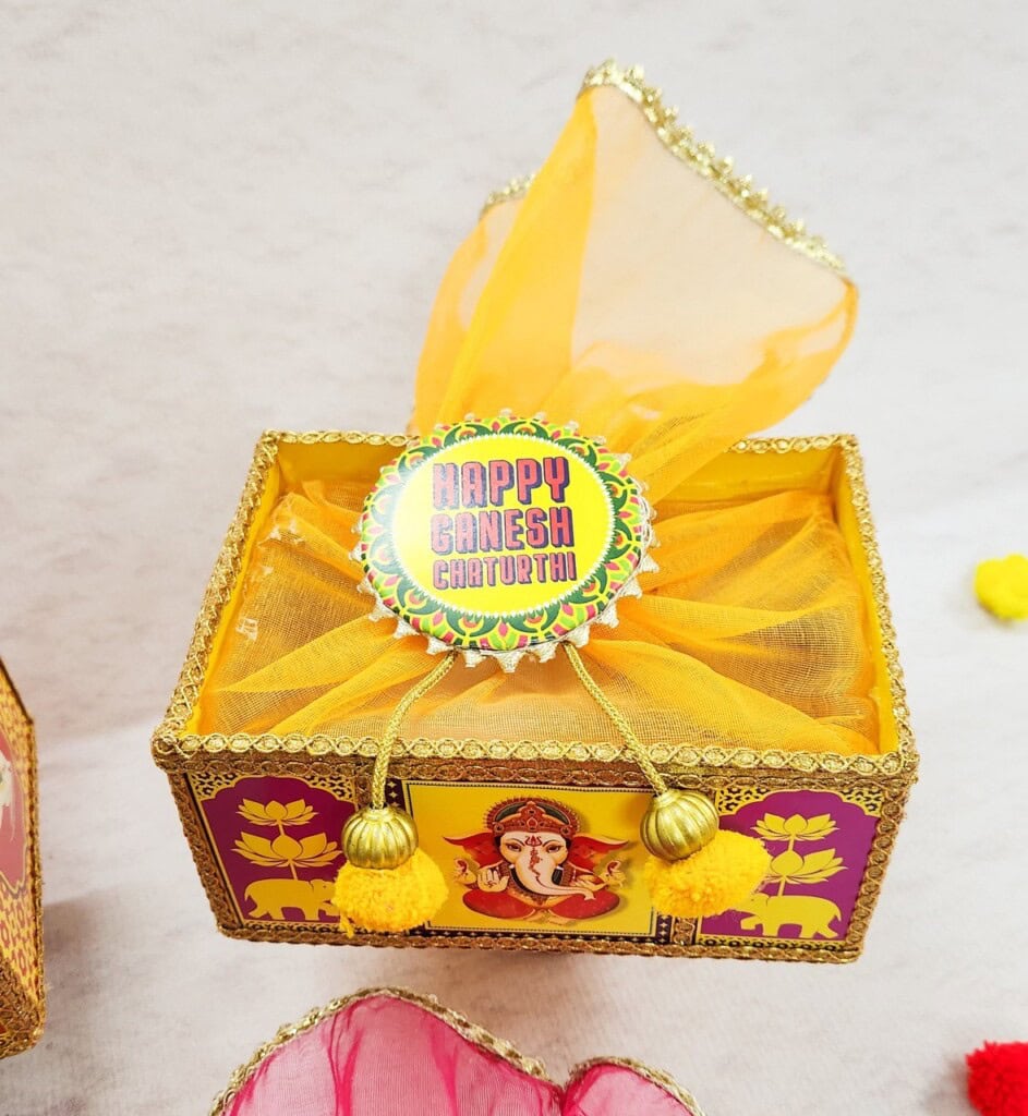 Floral Art presents A decorated box with an orange scarf and golden bells, featuring an illustration of Lord Ganesha and a "Happy Ganesh Chaturthi" sign on top.