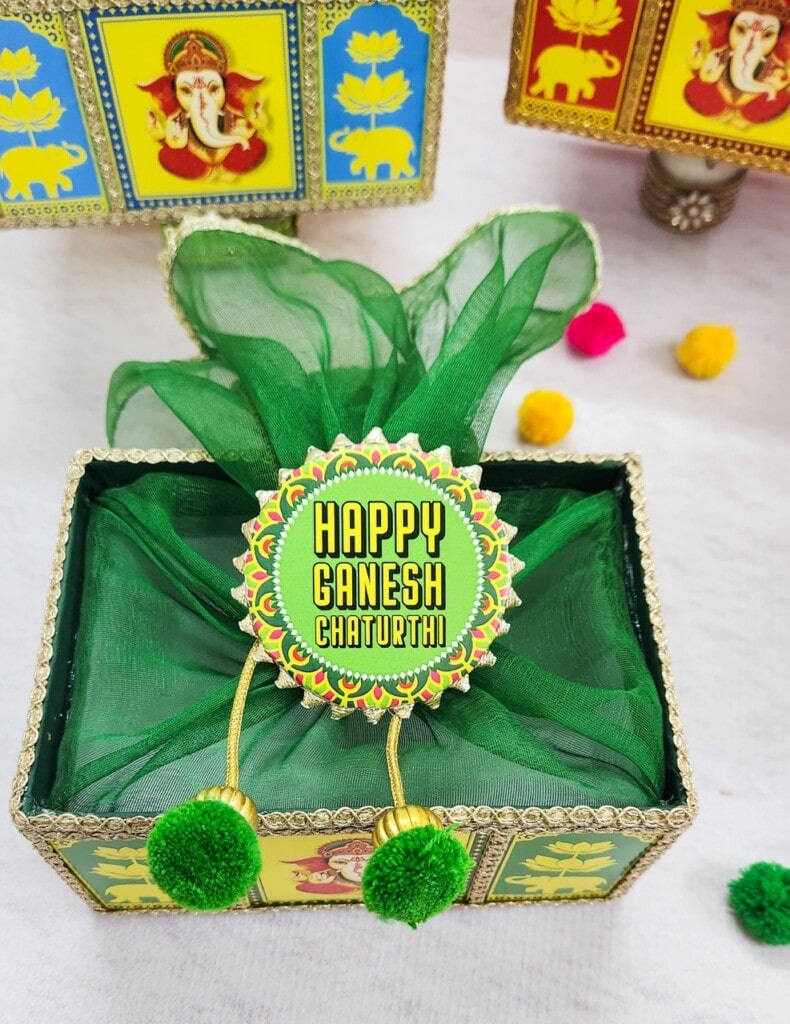 Floral Art presents A decorative box wrapped with green cloth and adorned with a "Happy Ganesh Chaturthi" badge with colorful motifs. Surrounding decorations include images of the deity Ganesh and small pom poms.