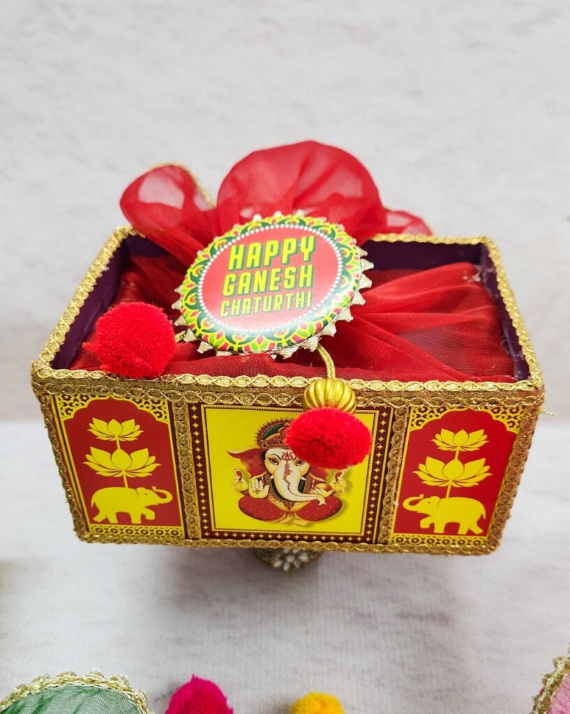 Floral Art presents A decorated box with an image of Lord Ganesha, adorned with red pom poms and embroidered edges. A badge on the box reads "Happy Ganesh Chaturthi!.